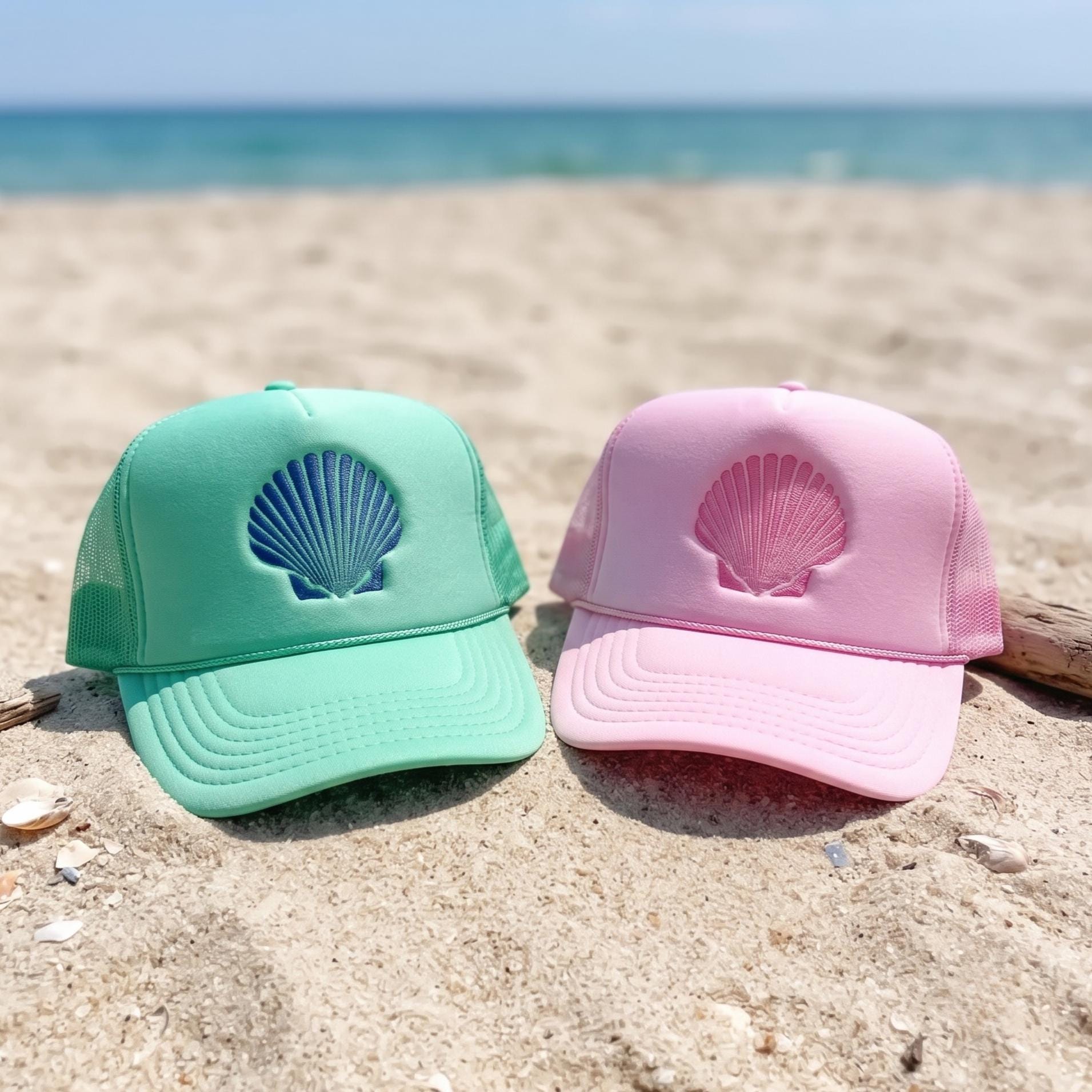 Mint and pink seashell embroidered trucker hats displayed on a sandy beach