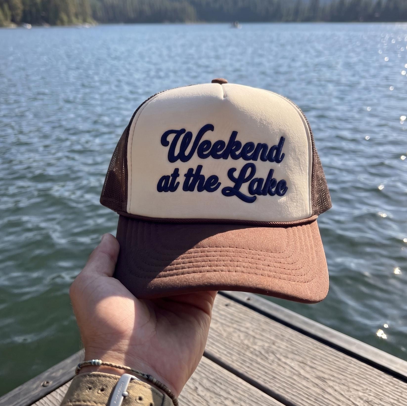 Hand holding a brown and cream Weekend at the Lake trucker hat by the water