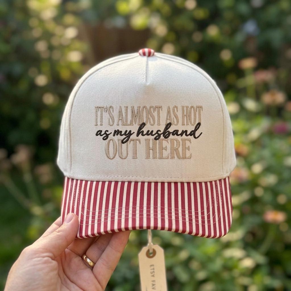 Hand holding a cream embroidered quote baseball cap with red striped brim outdoors
