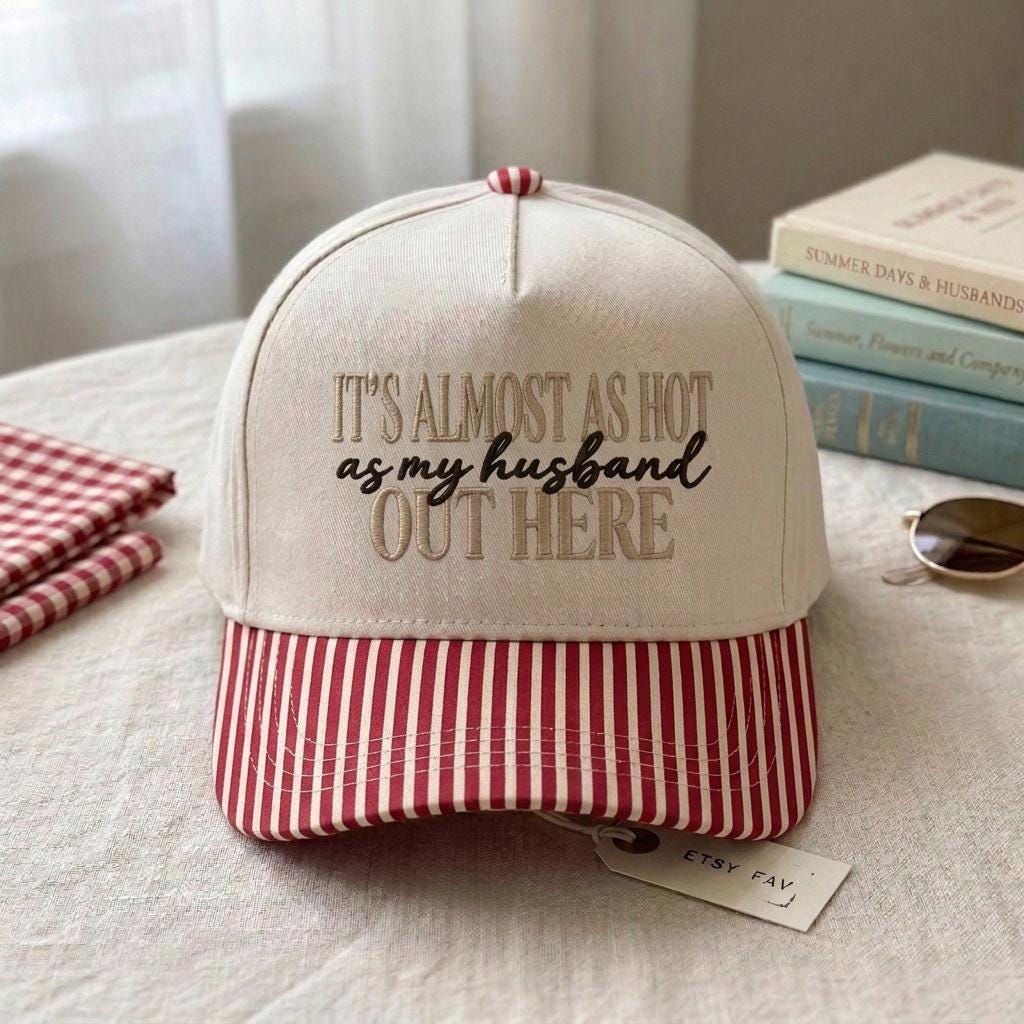Cream baseball cap with embroidered quote reading It’s Almost as Hot as My Husband Out Here and red striped brim