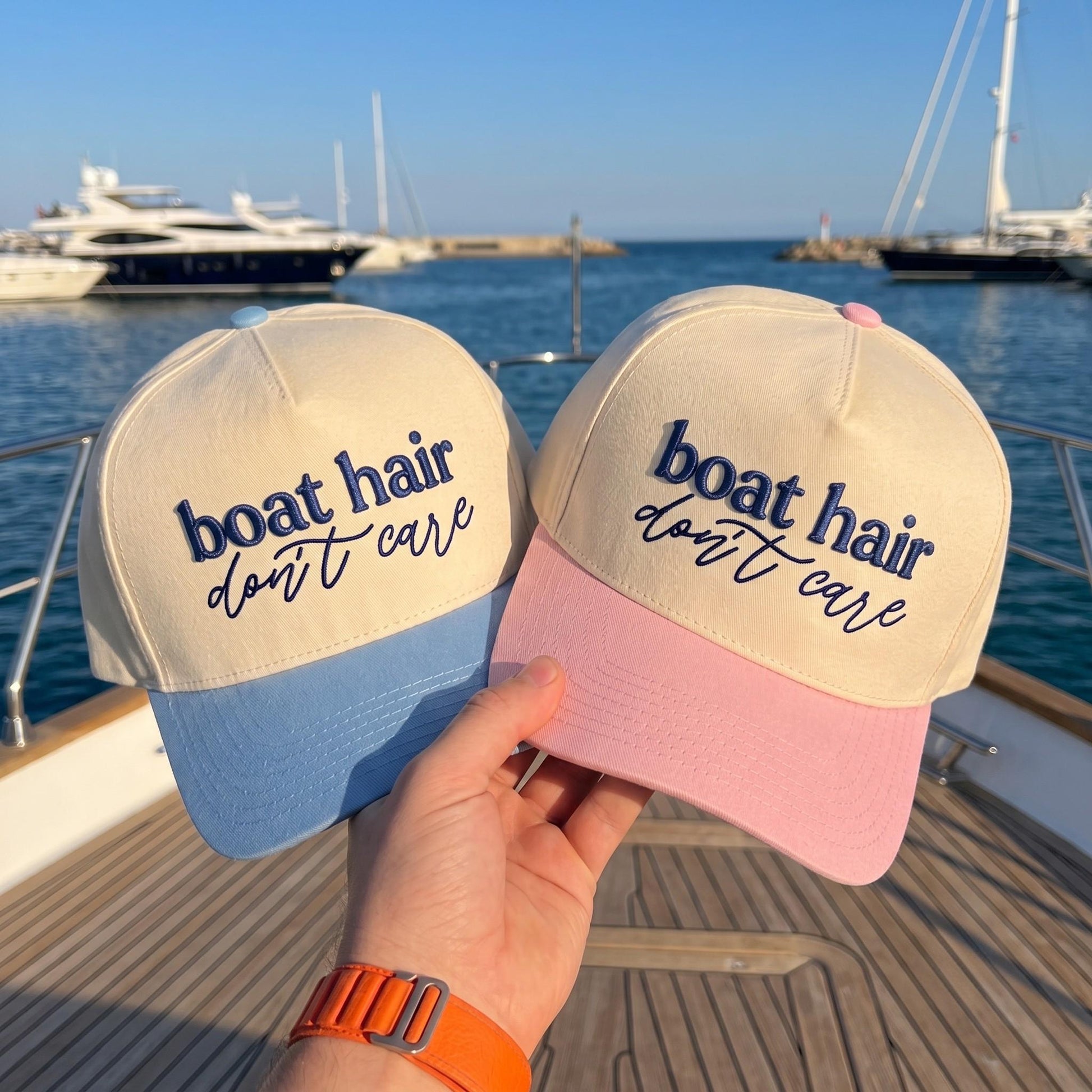 Two cream embroidered Boat Hair Don't Care hats with blue and pink brims by the water
