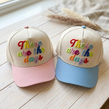 Pair of pastel two-tone caps displaying raised multicolor “These are the days” embroidery