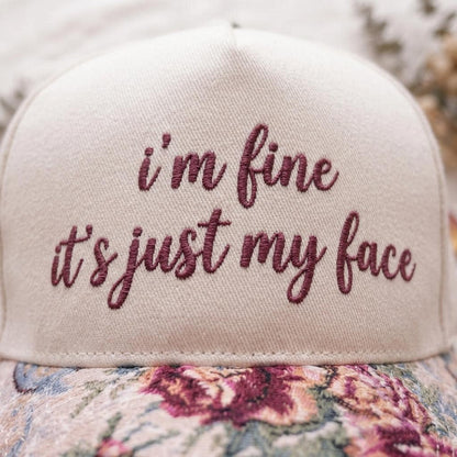 Close-up of burgundy script embroidery reading “I’m Fine, It’s Just My Face” on a hat