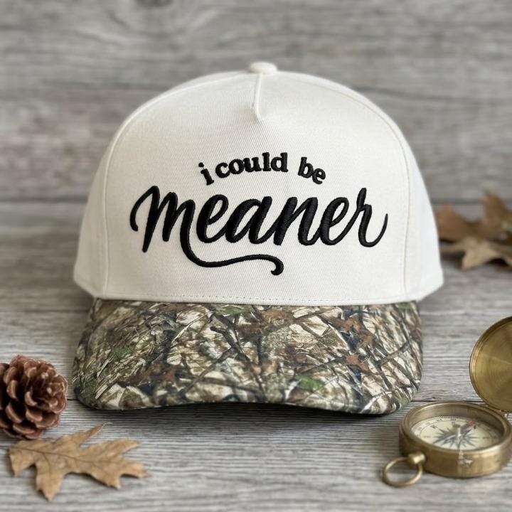 Embroidered “i could be meaner” camo brim hat displayed on a rustic table