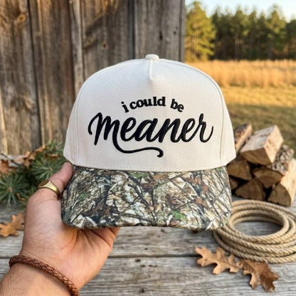 White embroidered hat with black “i could be meaner” lettering and camouflage brim outdoors