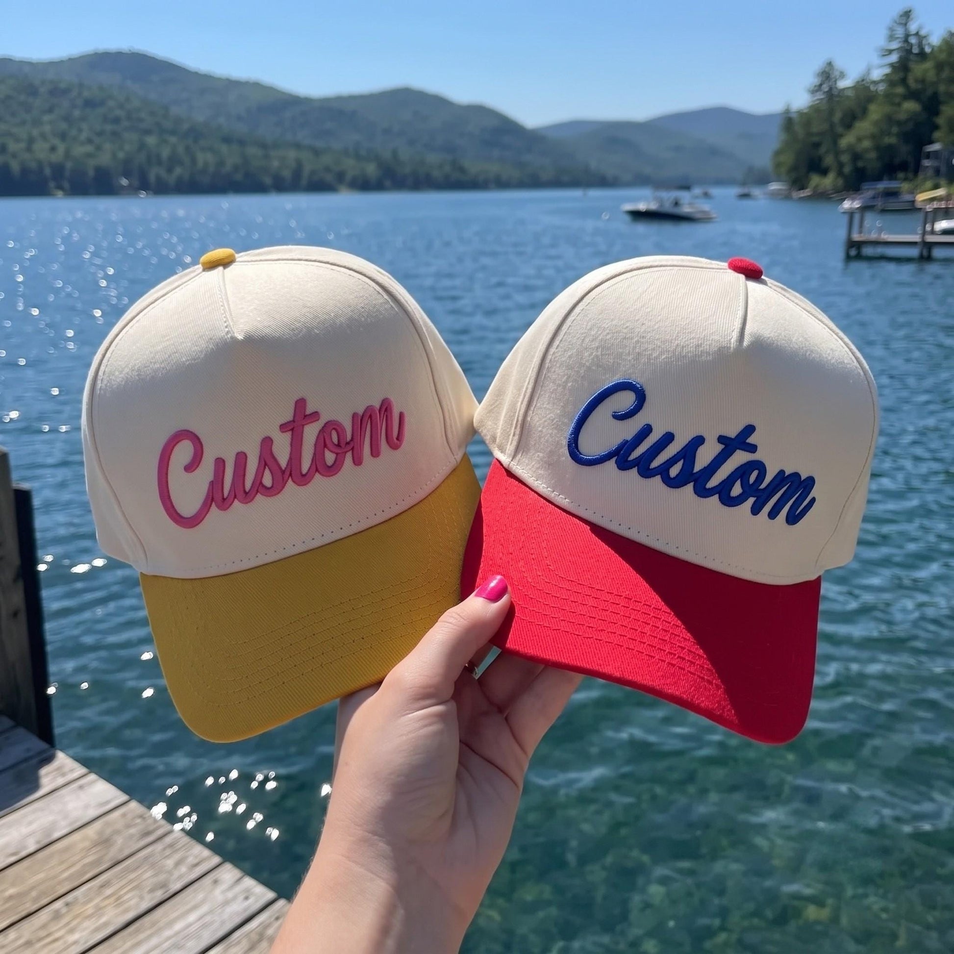 Two embroidered hats with script Custom lettering in pink and blue beside a lake