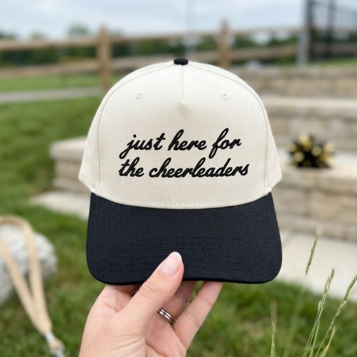 Two-tone embroidered hat with “just here for the cheerleaders” script shown outdoors