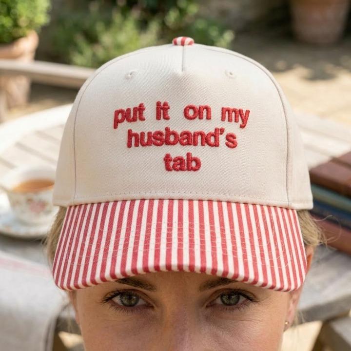 Person wearing a khaki baseball cap with red striped brim and humorous embroidered phrase