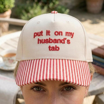 Person wearing a khaki baseball cap with red striped brim and humorous embroidered phrase