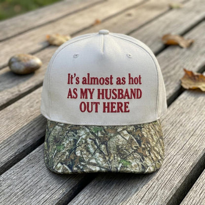 Funny embroidered hat with red text reading “It’s almost as hot as my husband out here” and a camouflage bill