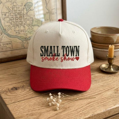 Front view of a cream and red Small Town Smoke Show embroidered trucker cap on a wooden table