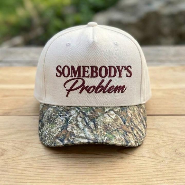 Cream embroidered hat with Somebody’s Problem lettering displayed on a wooden table
