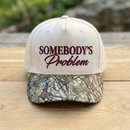Cream embroidered hat with Somebody’s Problem lettering displayed on a wooden table