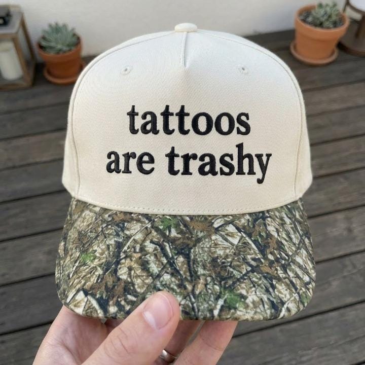Front view of cream embroidered hat with “tattoos are trashy” text and camouflage-patterned brim