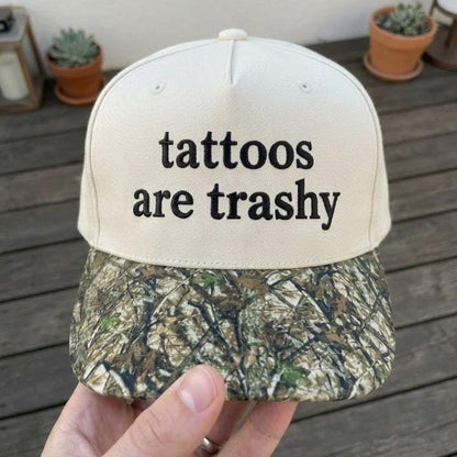 Front view of cream embroidered hat with “tattoos are trashy” text and camouflage-patterned brim