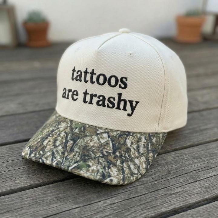 Angled view of embroidered “tattoos are trashy” hat with camouflage-patterned brim