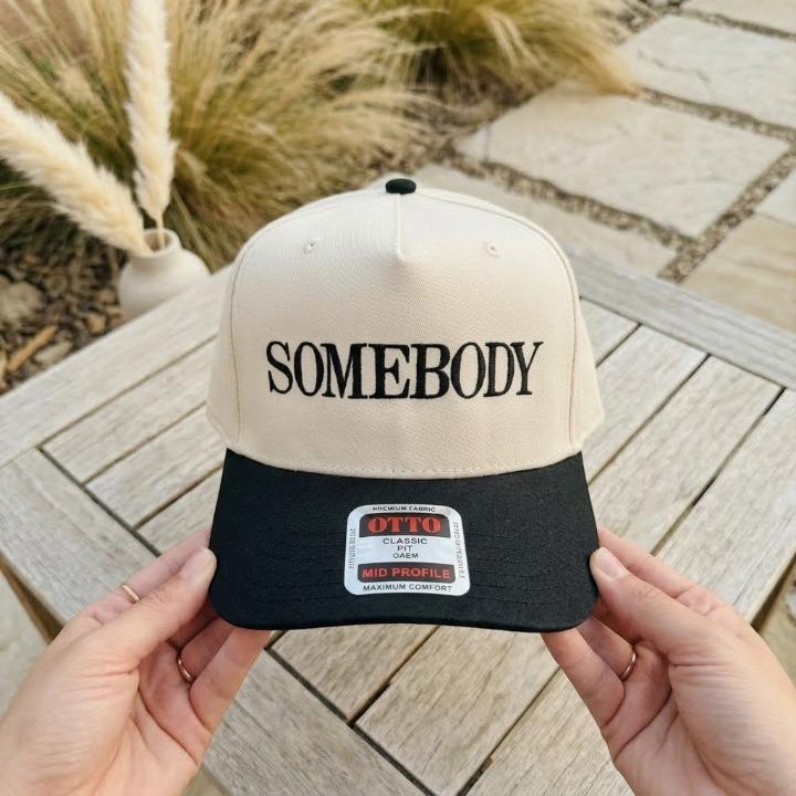 Close-up of a cream and black trucker hat embroidered with Somebody