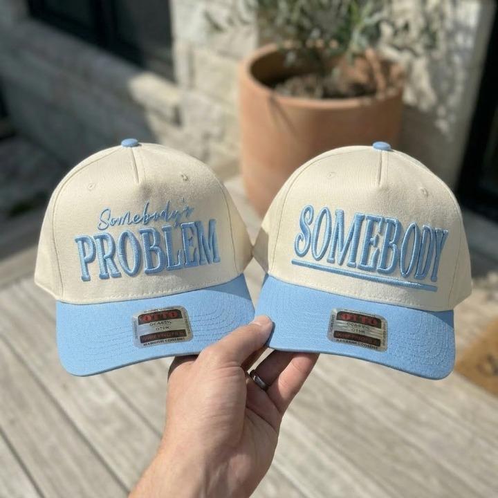 Two cream and light blue embroidered trucker hats reading Somebody’s Problem and Somebody