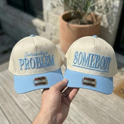 Two cream and light blue embroidered trucker hats reading Somebody’s Problem and Somebody