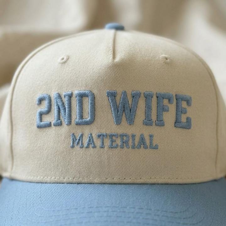 Close-up of blue embroidered 2ND WIFE MATERIAL lettering on a cream hat