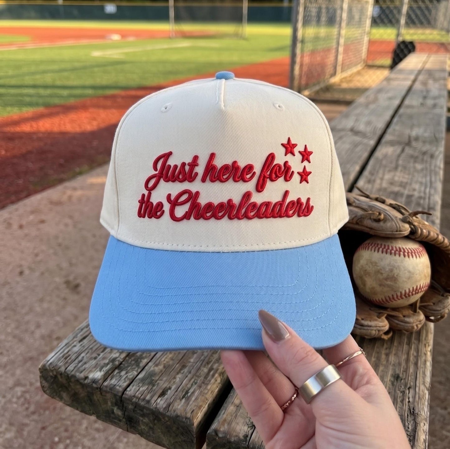 Embroidered cheerleader hat with red script lettering, star accents, and a light blue brim at a baseball field