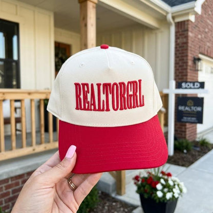 Two-tone REALTORGRL cap with cream crown and red bill held in front of a house