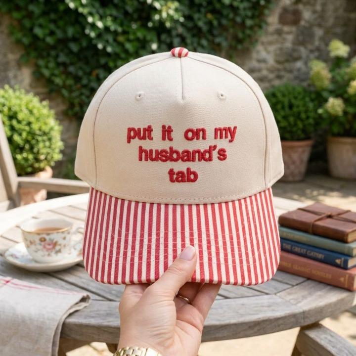 Khaki embroidered baseball cap with red striped brim and “put it on my husband’s tab” text