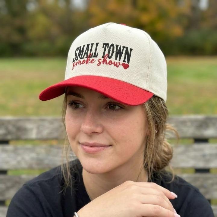 Person wearing a cream and red Small Town Smoke Show embroidered trucker cap outdoors