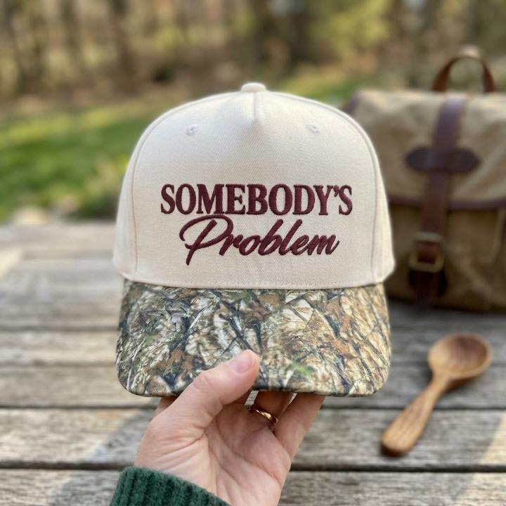 Front view of cream hat with maroon Somebody’s Problem embroidery and camouflage-pattern bill