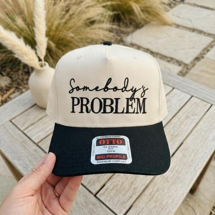 Close-up of a cream and black trucker hat embroidered with Somebody’s Problem