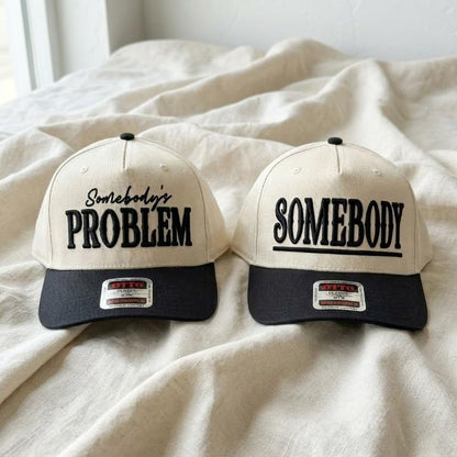 Two cream and black embroidered trucker hats with Somebody’s Problem and Somebody lettering